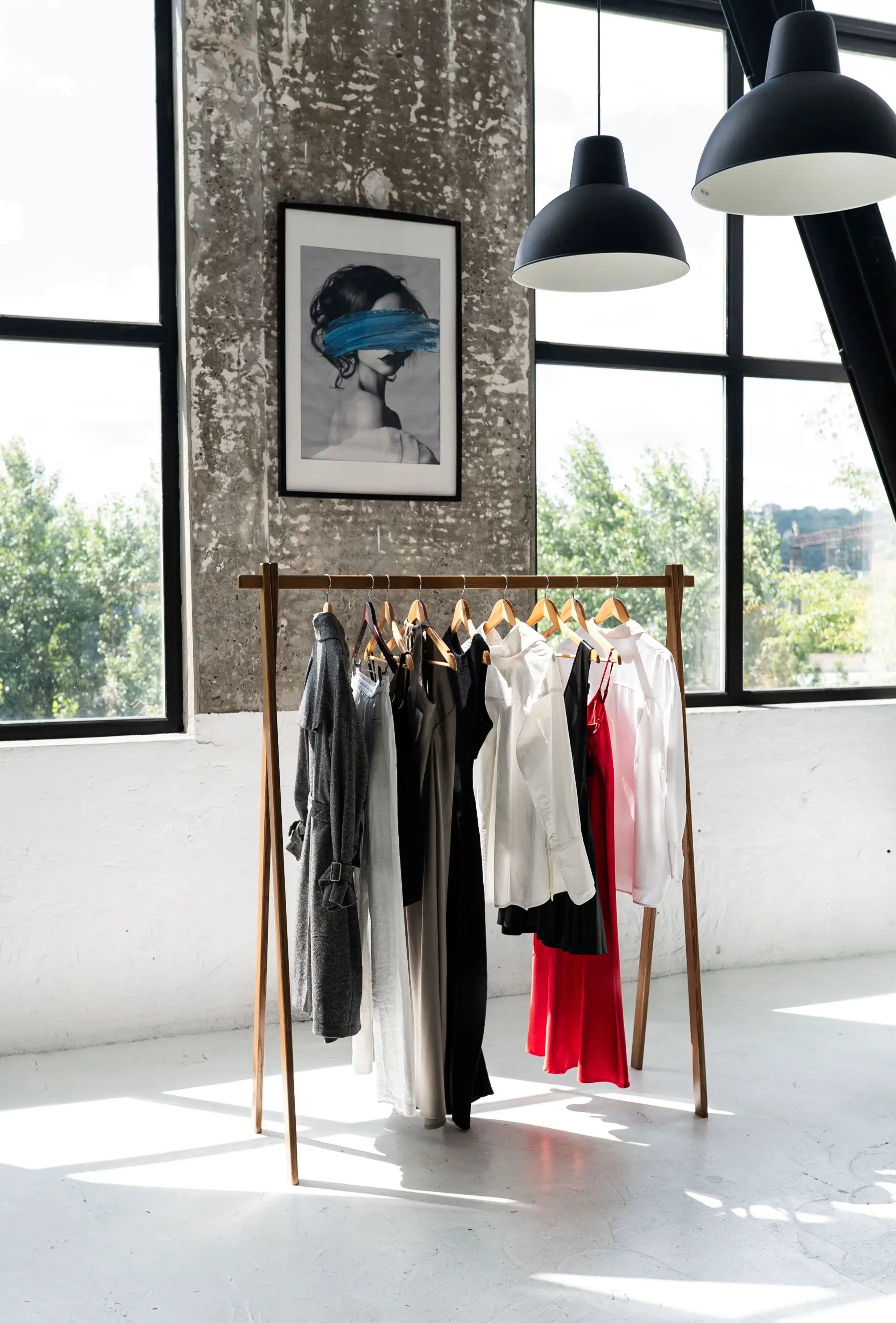 Clothing rack with clothes in a room with large windows and a framed picture on the wall.