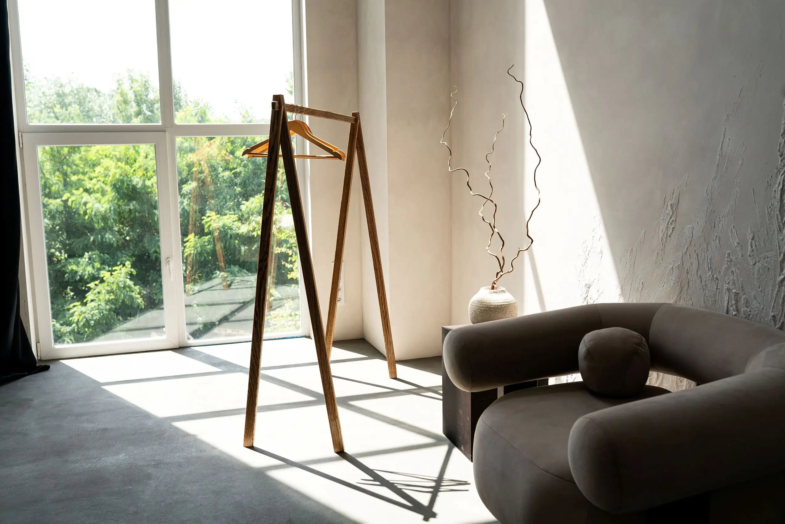 Modern interior with a wooden coat rack, gray sofa, and large window with greenery outside.