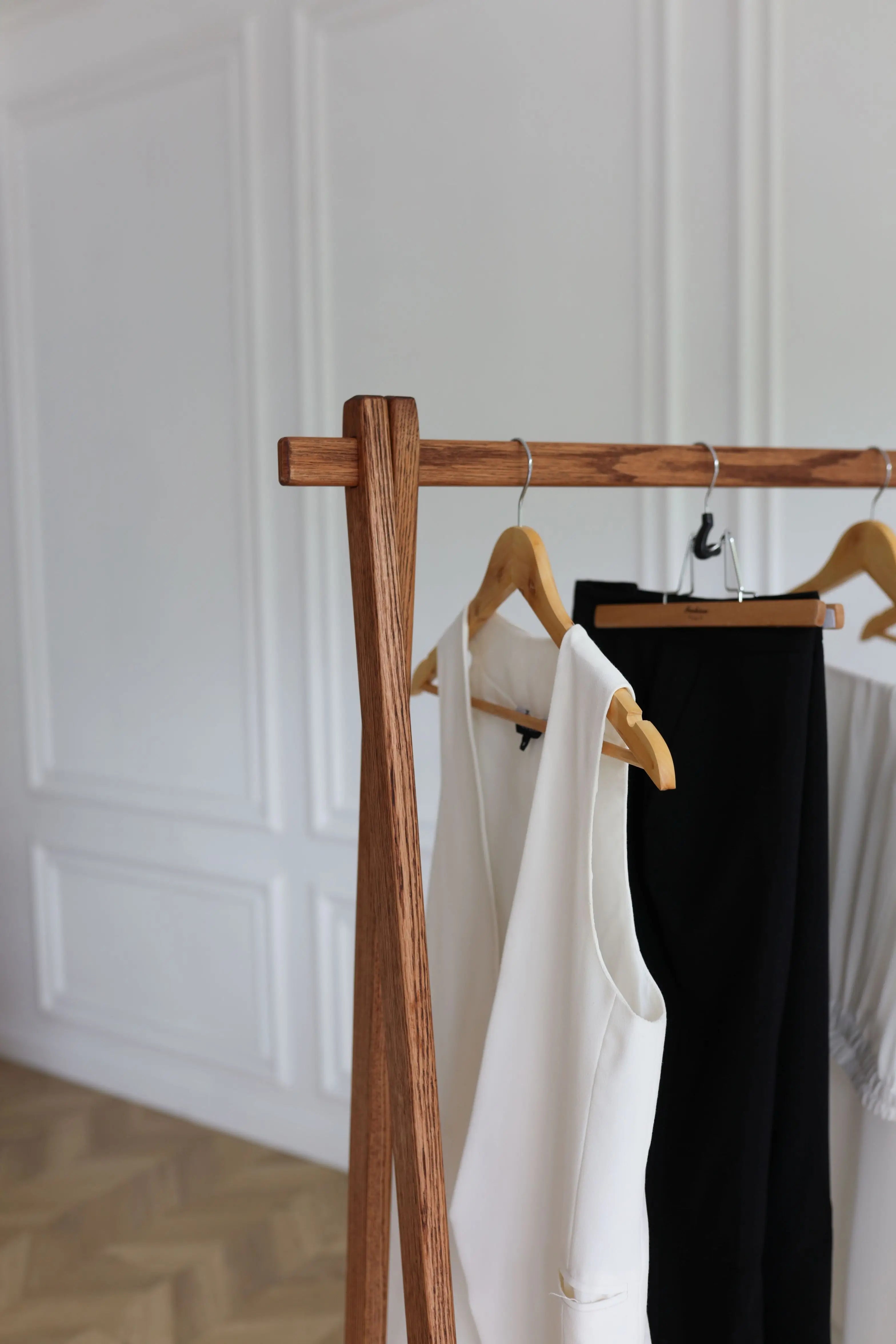 Wooden clothing rack with a white garment and black pants against a white paneled wall.