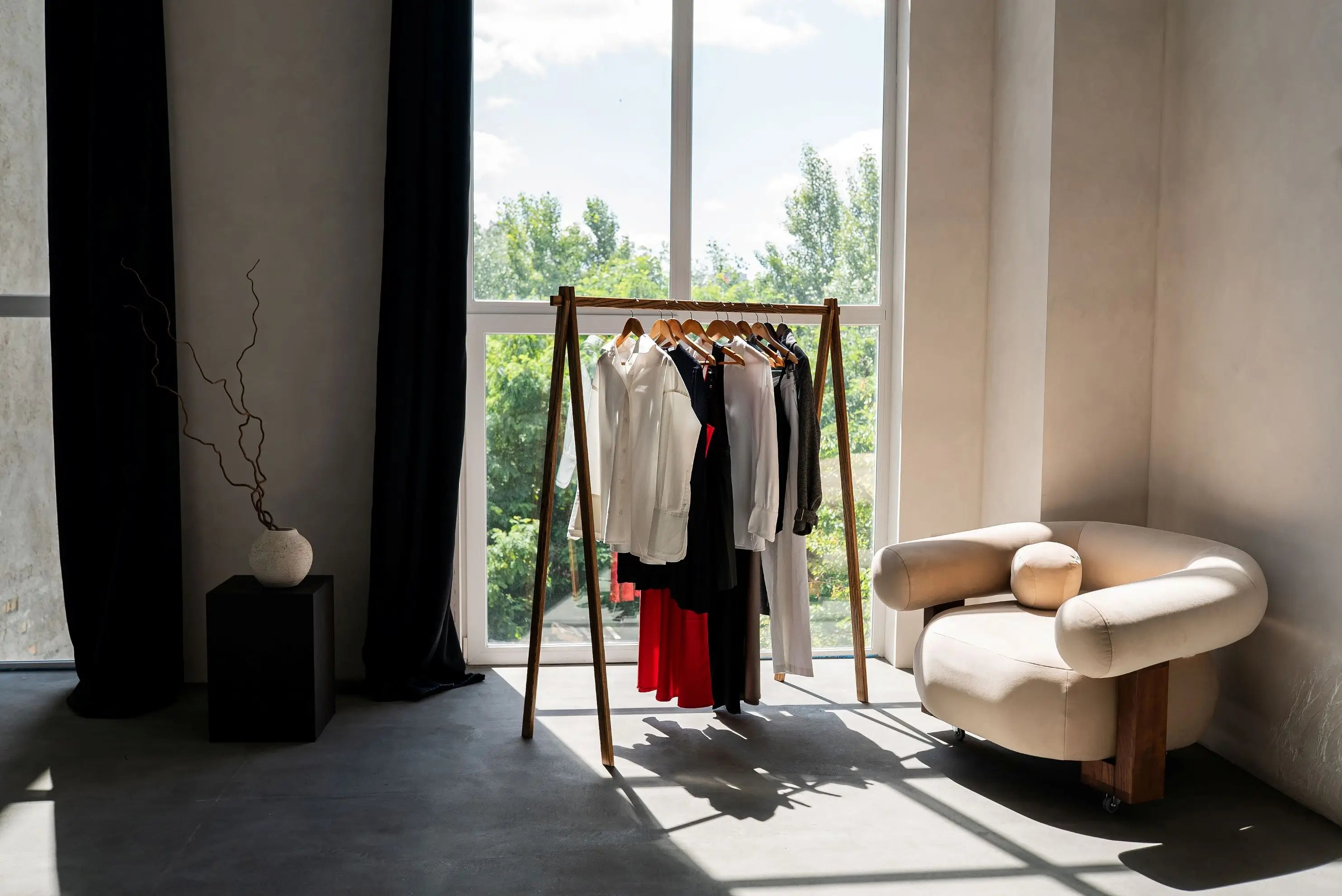 Room with a clothing rack against a window, featuring a modern armchair.