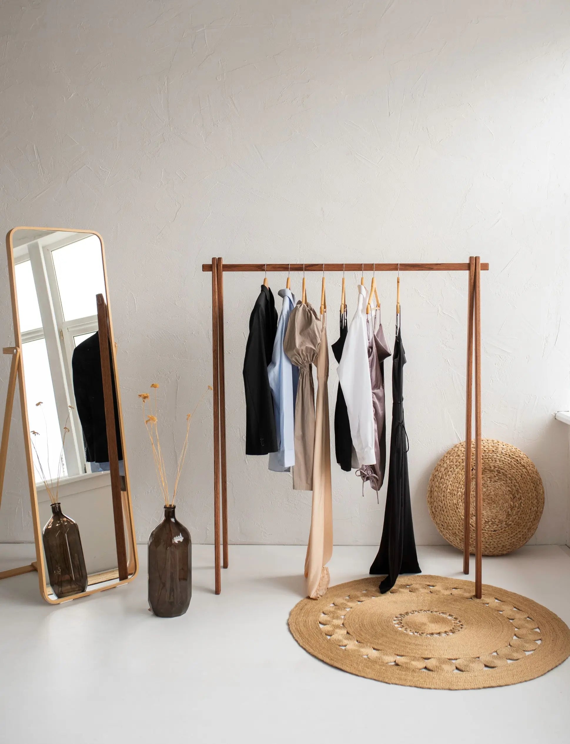 Wooden clothing rack with hanging clothes next to a standing mirror and decorative items on a light floor.