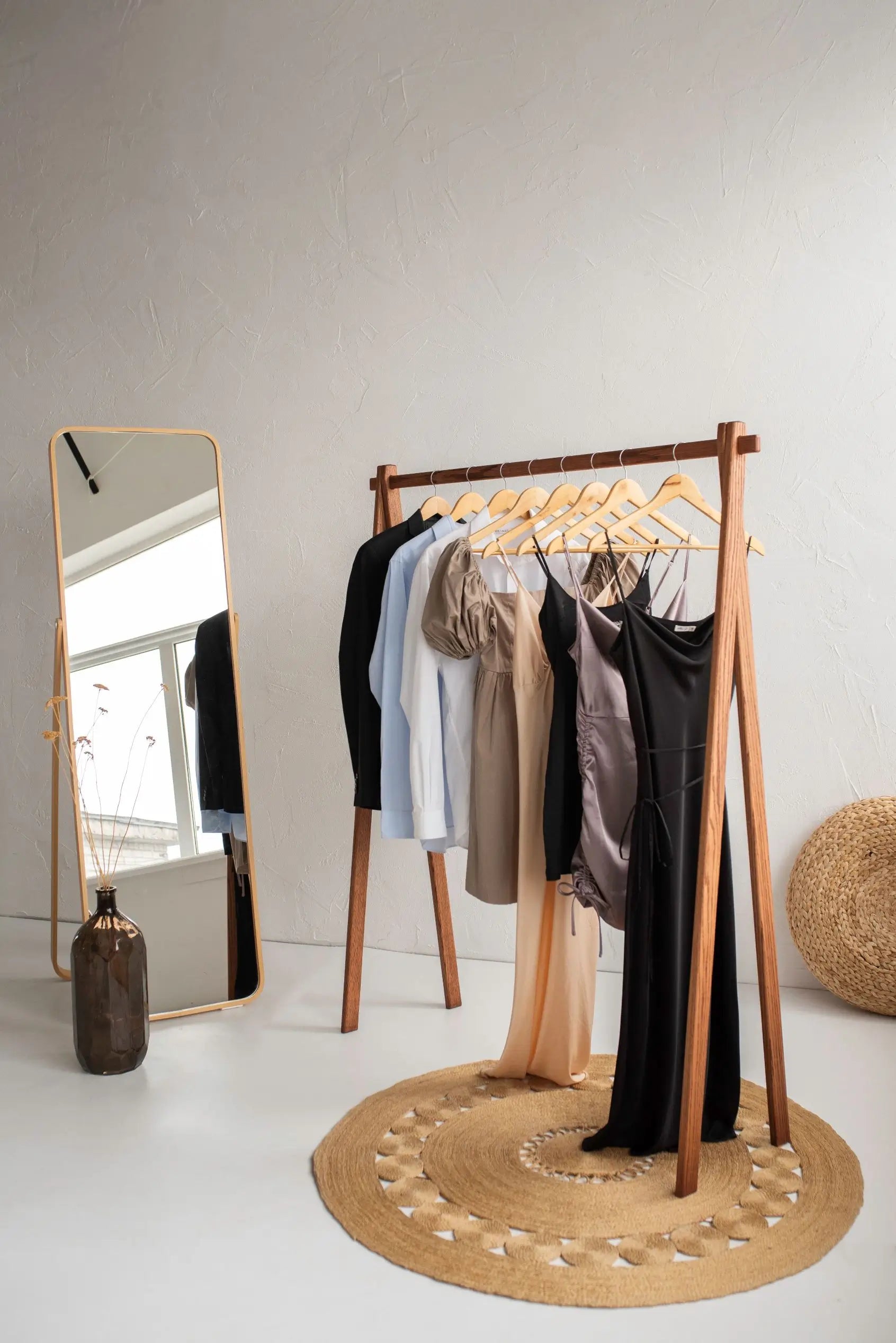 Wooden clothing rack with hangers holding clothes next to a full-length mirror on a white floor.